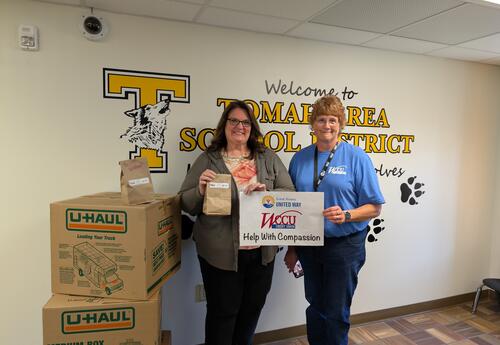 two women stand next to a stack of brown boxes and hold brown paper snack bags and a sign that says WCCU