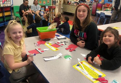 An older student works with a couple of younger students on an art project. They are seated at a table with art supplies in front of them. They are smiling at the camera.