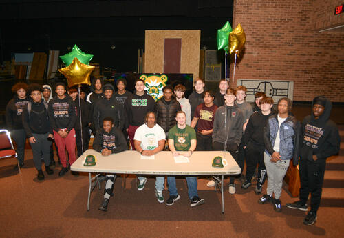 College signees sit at a table surrounded by teammates