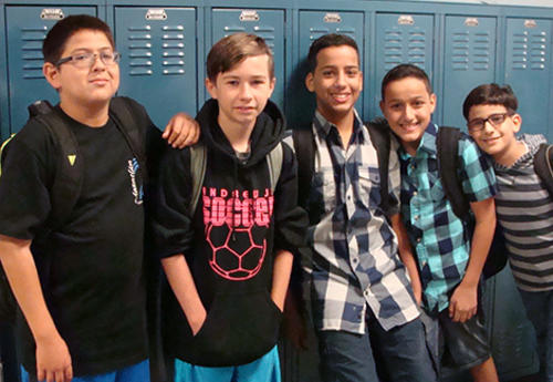 Male students pose against a row of blue lockers, wearing backpacks and smiling.