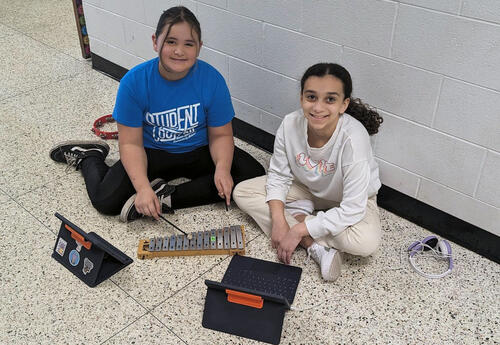 Two fifth grade girls smile in the hallway while playing with instruments and ipads