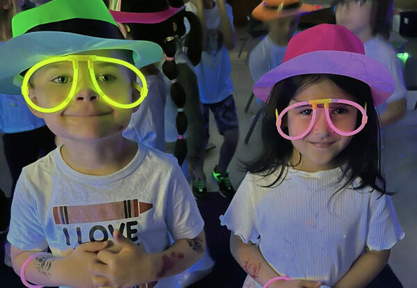 Boy and girl kindergarten students smile while wearing glow in the dark hats and glasses for glow party