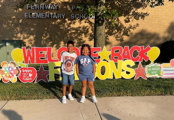 Two students pose proudly in front of welcome back sign in front of school