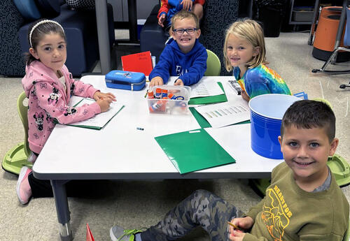 Four young students sitting at a classroom table and smiling.