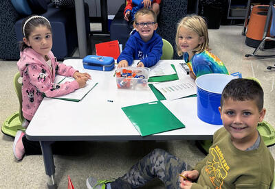 Four young students sitting at a classroom table and smiling.
