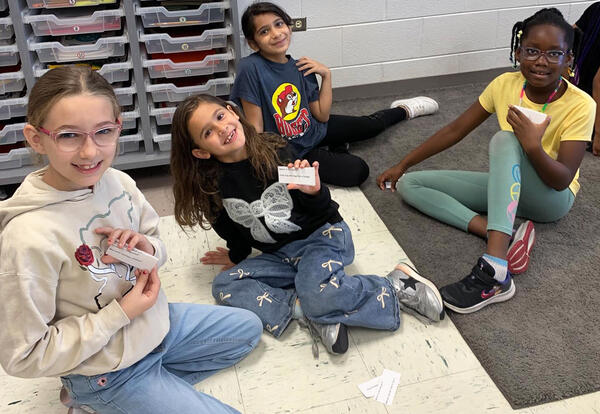 Four elementary students sitting on the floor in a classroom, smiling and holding cards during an activity