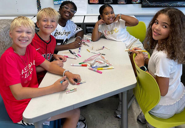 Five smiling children sit around a classroom table, coloring