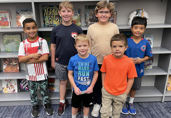 : Six young boys stand together in a library, smiling for the camera.