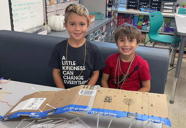 Two students smile while sitting at a classroom table covered with craft supplies and cardboard, building bridges during STEM club