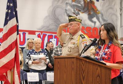 Students speaking at podium while veteran salutes in front of crowd