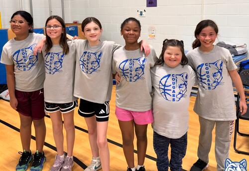 Elementary girls stand arm in arm in a gymnasium wearing matching basketball team shirts and smiling at the camera.