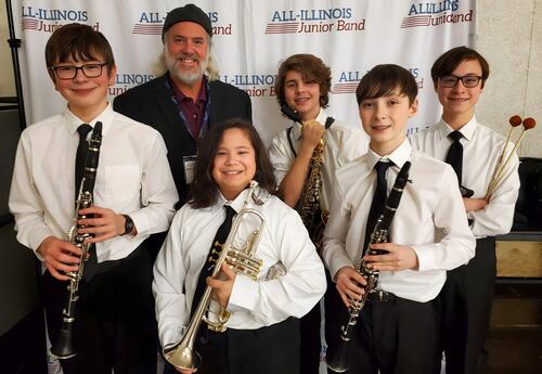 Middle school band students and their director pose with instruments in front of an All-Illinois Junior Band backdrop.