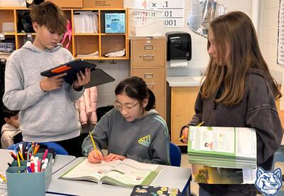 Two students standing next to another student sitting at desk working together in classroom