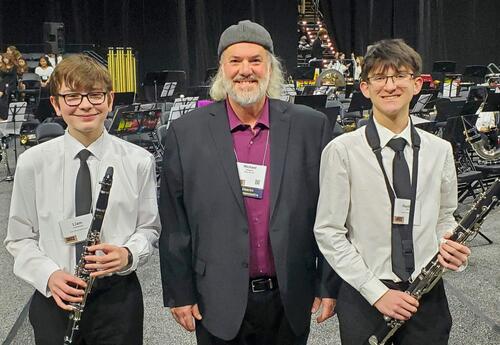 Band director and two young musicians holding clarinets stand in front of an orchestra on stage.