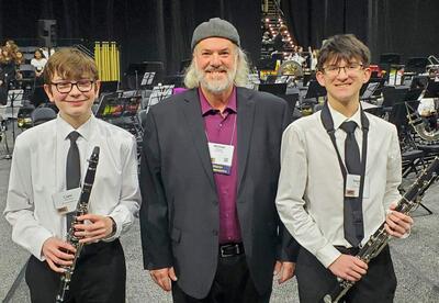 Band director and two young musicians holding clarinets stand in front of an orchestra on stage.