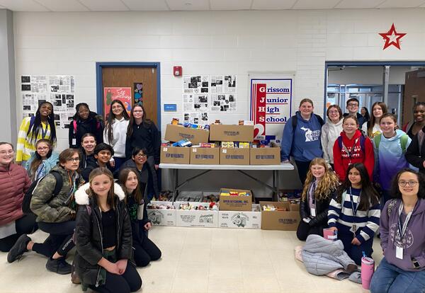 A group of students pose in front of boxes of food donations with a Grissom Junior High School sign visible.