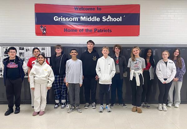 A group of students stands in front of lockers with a banner that reads "Welcome to Grissom Middle School Home of the Patriots".
