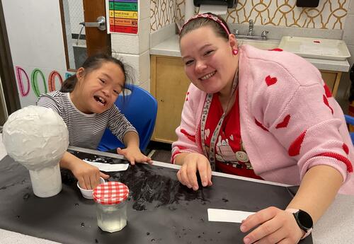 Child and teacher working together on a project and smiling at a table.
