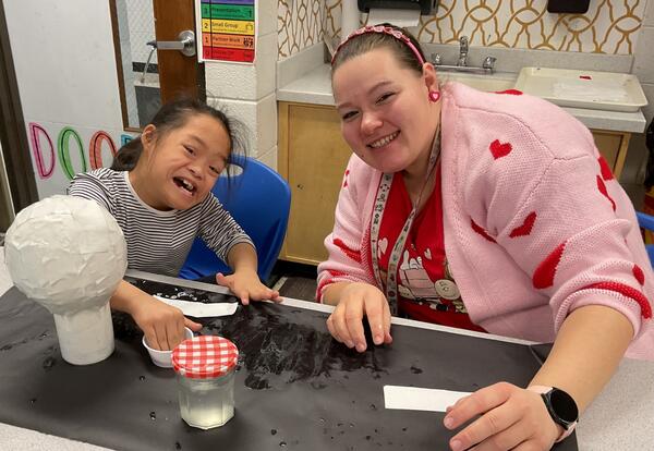Child and teacher working together on a project and smiling at a table.