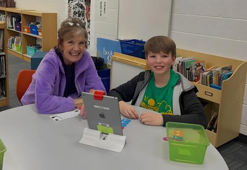 A smiling woman and child sit at a round table with a tablet and art supplies in a library or classroom setting.
