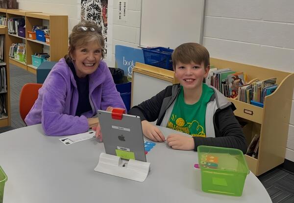 A smiling woman and child sit at a round table with a tablet and art supplies in a library or classroom setting.