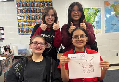 Four students pose for a photo in a classroom, two giving thumbs up while one holds a drawing of a house labeled "House".