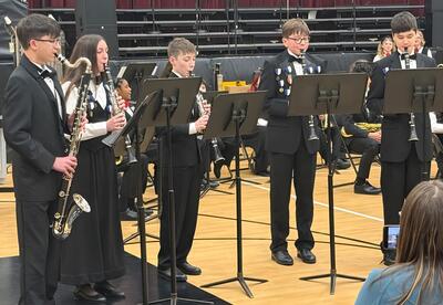 A group of musicians in formal attire playing woodwind instruments in a school gymnasium.