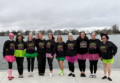 Individuals wearing matching black sweatshirts and colorful tutus pose proudly on a walkway by a lake at the Polar Plunge