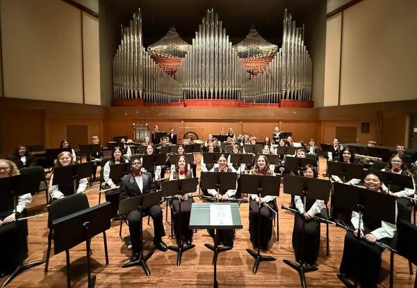 A concert band is seated on a stage in front of a large pipe organ, with music stands in front of them. Many members are holding instruments and wearing formal attire.