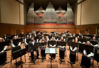 A concert band is seated on a stage in front of a large pipe organ, with music stands in front of them. Many members are holding instruments and wearing formal attire.