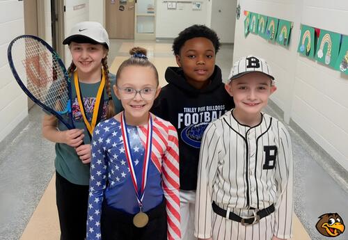 Four children stand in a hallway. One holds a tennis racket, one wears a baseball uniform, one wears a football hoodie, and one wears a patriotic-themed outfit.