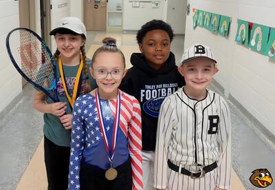 Four children stand in a hallway. One holds a tennis racket, one wears a baseball uniform, one wears a football hoodie, and one wears a patriotic-themed outfit.
