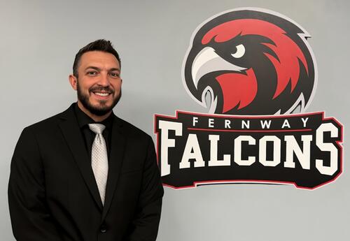 New Fernway school principal smiles next to Fernway Falcons logo in board room