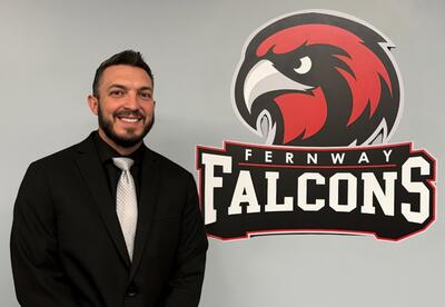 New Fernway school principal smiles next to Fernway Falcons logo in board room