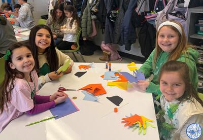 Four children sit at a table crafting with colorful paper, scissors, and glue sticks.