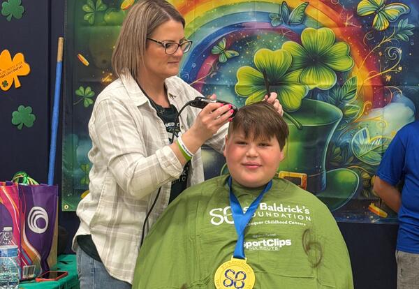A person uses clippers to shave the hair of a child wearing a green cape and a medal for St. Baldrick's.