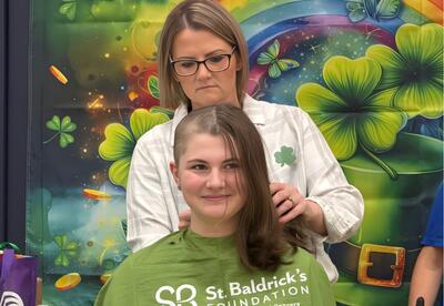 A student with half their head shaved sits with a green cape draped over them, while a hair stylist cuts the other half for St. Baldrick's.