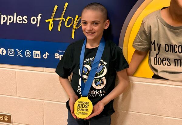 A student with a shaved head proudly displays a gold medal around their neck at St. Baldrick's event.