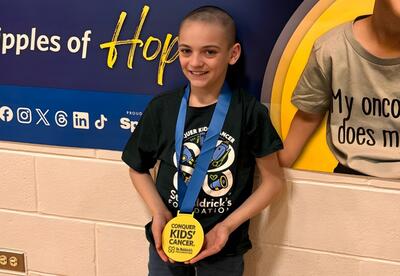 A student with a shaved head proudly displays a gold medal around their neck at St. Baldrick's event.