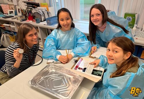 A teacher and three students in blue gowns are gathered around a table with a stethoscope, papers, and a foil tray in an operating room project.