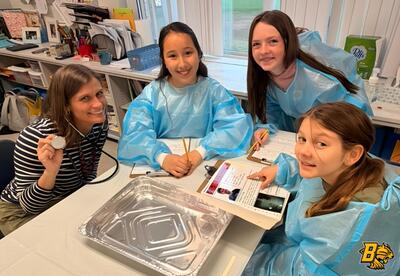 A teacher and three students in blue gowns are gathered around a table with a stethoscope, papers, and a foil tray in an operating room project.