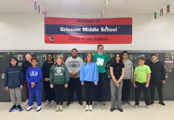 A group of students stands in front of lockers with a banner that reads "Welcome to Grissom Middle School Home of the Patriots".