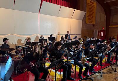 A jazz band performs in a gymnasium, with members playing saxophones, guitars, drums, and keyboard.