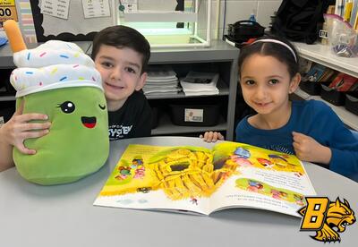 A child holds a large green plush toy while another child looks at an open book with colorful illustrations.