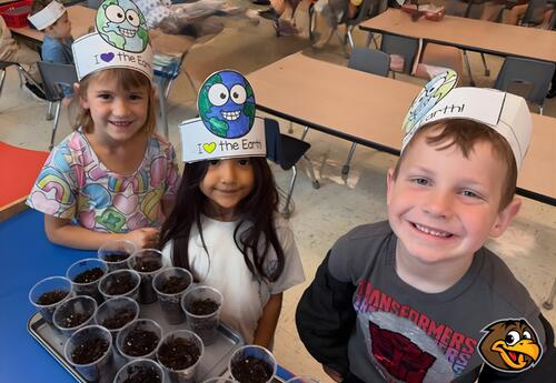 Three children wearing paper hats decorated with the Earth and the phrase "I love the Earth" smile with a tray of small cups filled with soil in front of them.