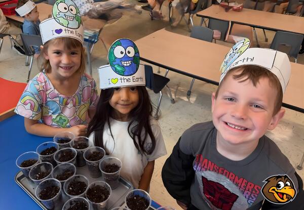 Three children wearing paper hats decorated with the Earth and the phrase "I love the Earth" smile with a tray of small cups filled with soil in front of them.