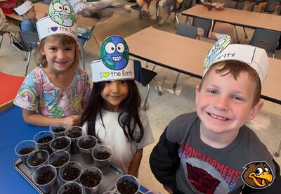 Three children wearing paper hats decorated with the Earth and the phrase "I love the Earth" smile with a tray of small cups filled with soil in front of them.