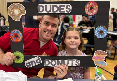 A student and guest smile through a photo frame decorated with donuts and the words "Dudes and Donuts."