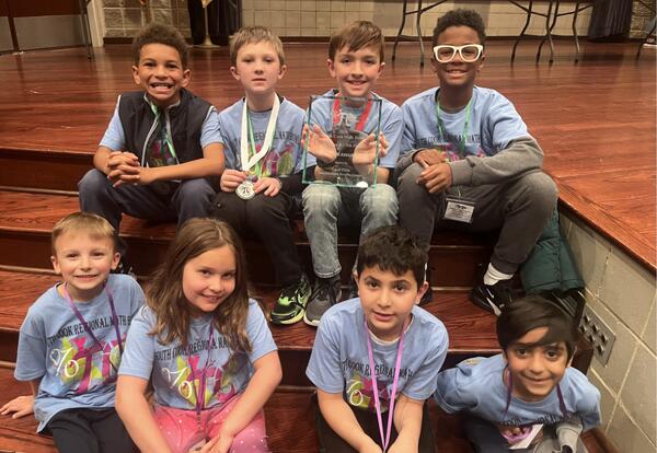 A group of children are sitting on wooden steps, smiling and wearing light blue t-shirts and medals. One child in the back row holds a glass trophy.