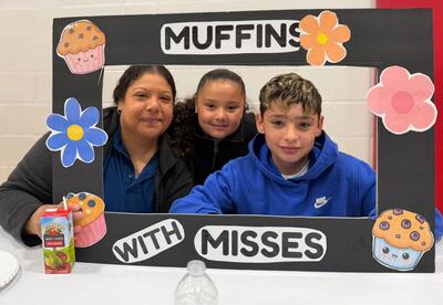 Three people pose behind a photo booth frame decorated with muffins and flowers. The frame reads "MUFFINS WITH MISSES".